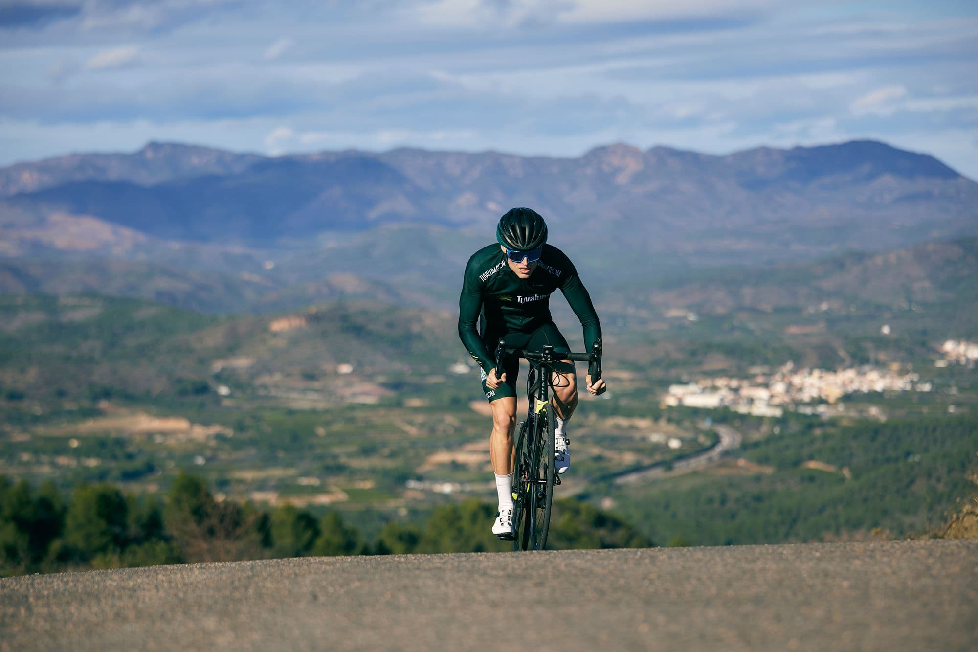 A cyclist climbing out of the saddle on a mountain road above a wide hazy valley